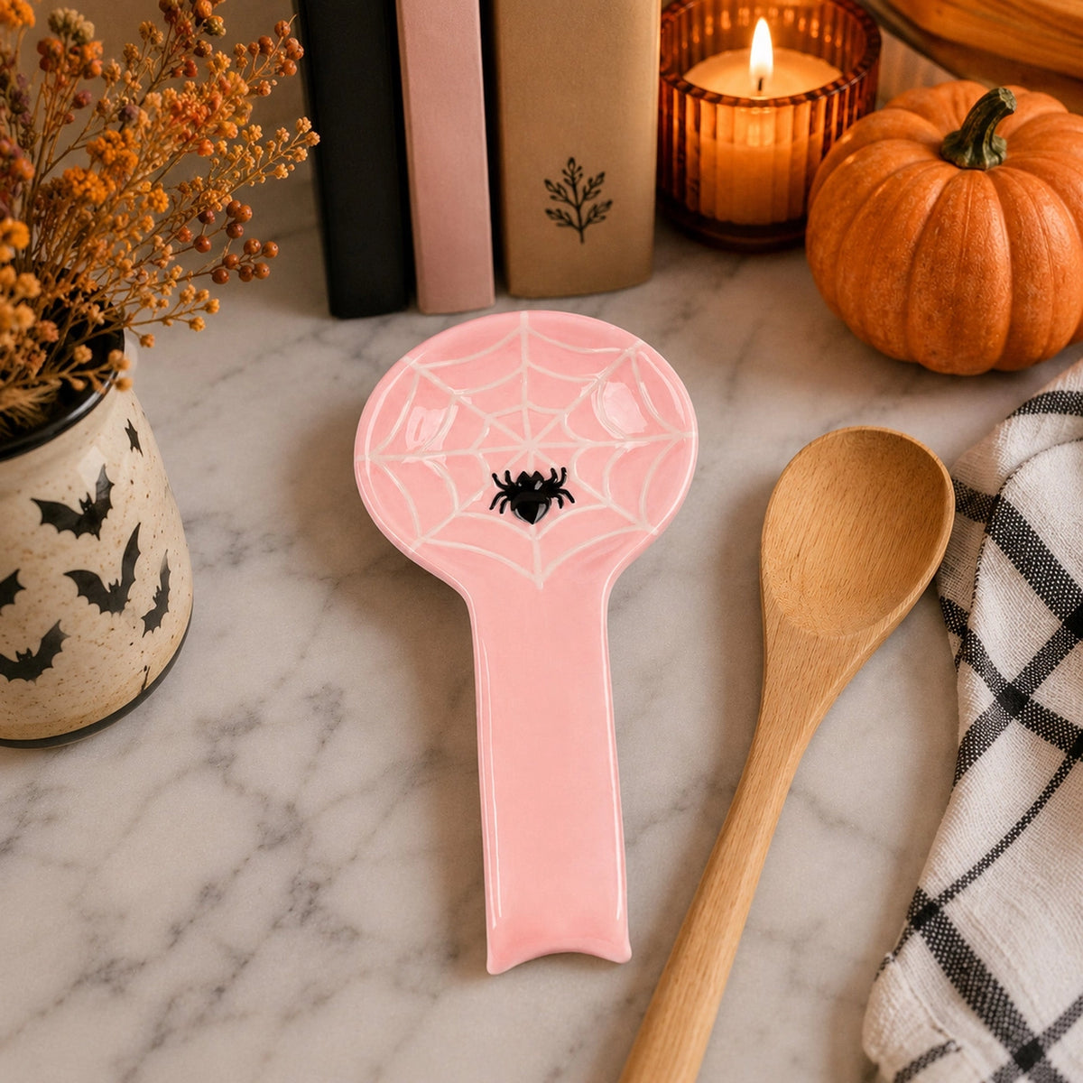 Pink spiderweb spoon rest beside a wooden spoon on a pale counter, with a pumpkin, books and a lit candle.