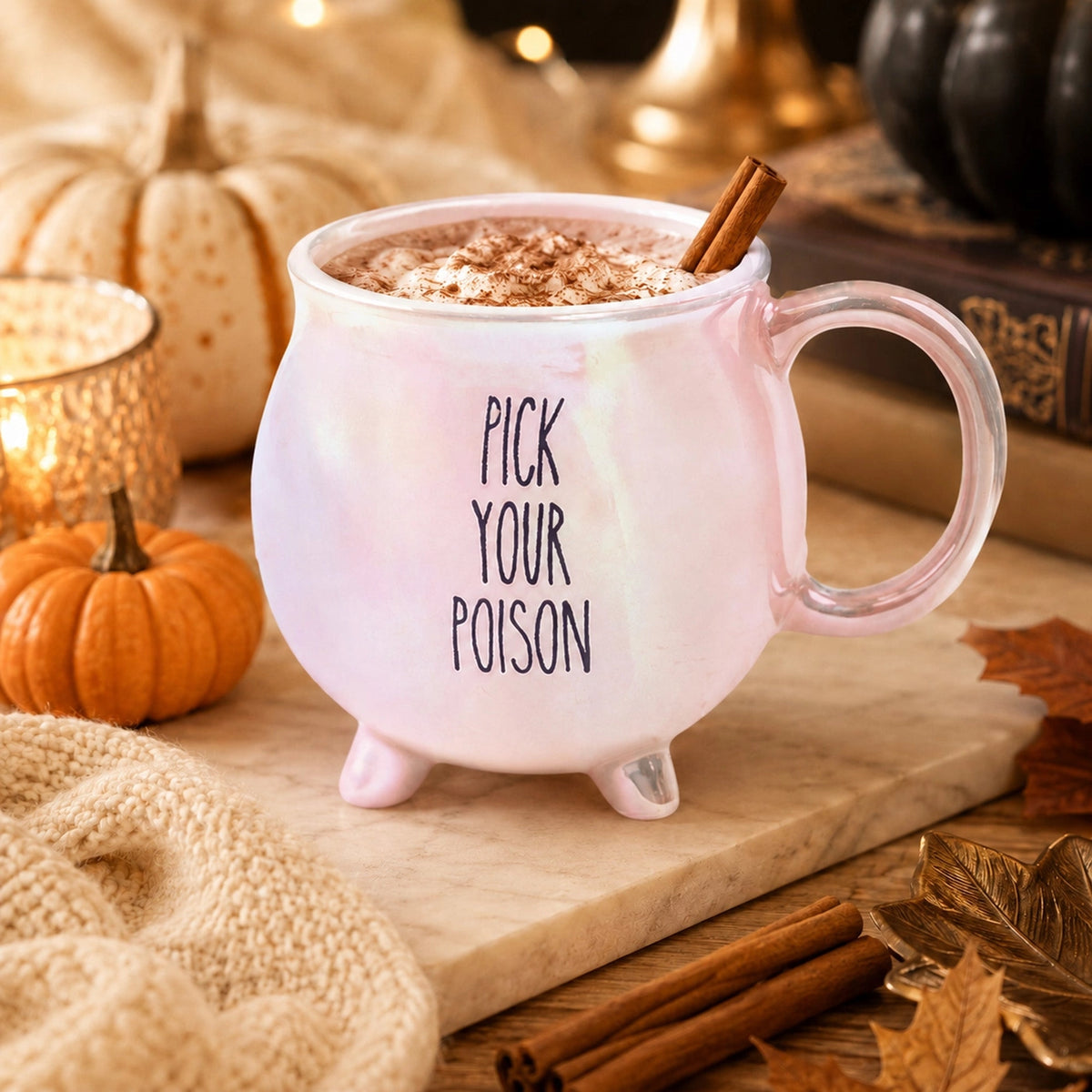 Pink iridescent Pick Your Poison cauldron mug with a cinnamon stick beside pumpkins and a candle.