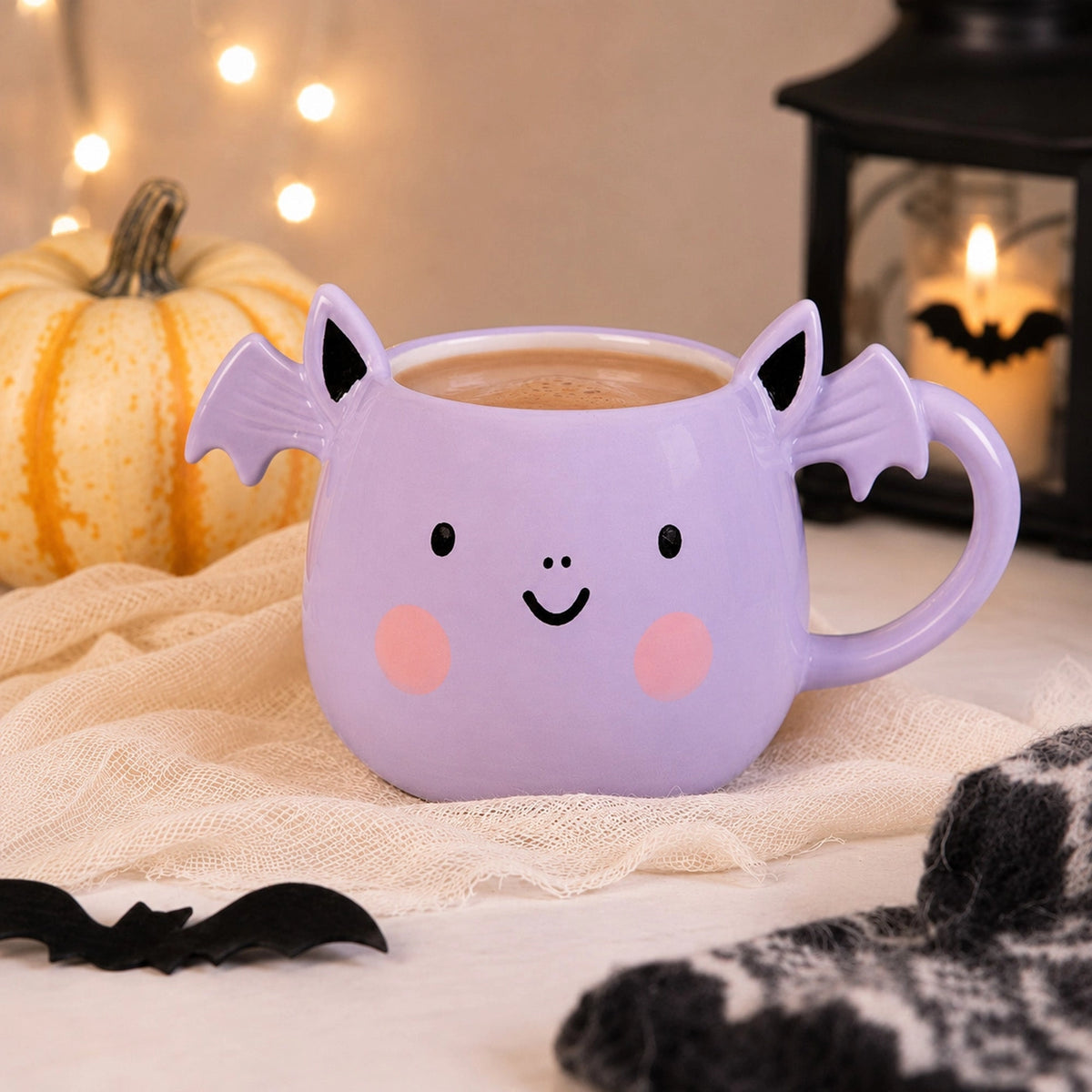 Smiling lilac bat mug with scalloped wings and pink cheeks beside a pale pumpkin and a glowing bat lantern.