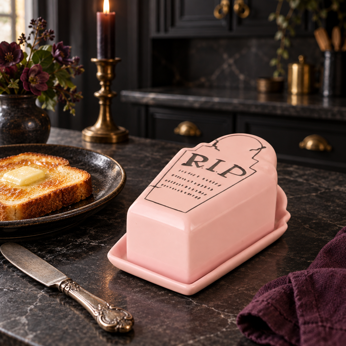 Pink R.I.P. butter dish beside toast and a plum cloth on a dark counter with a brass candleholder.