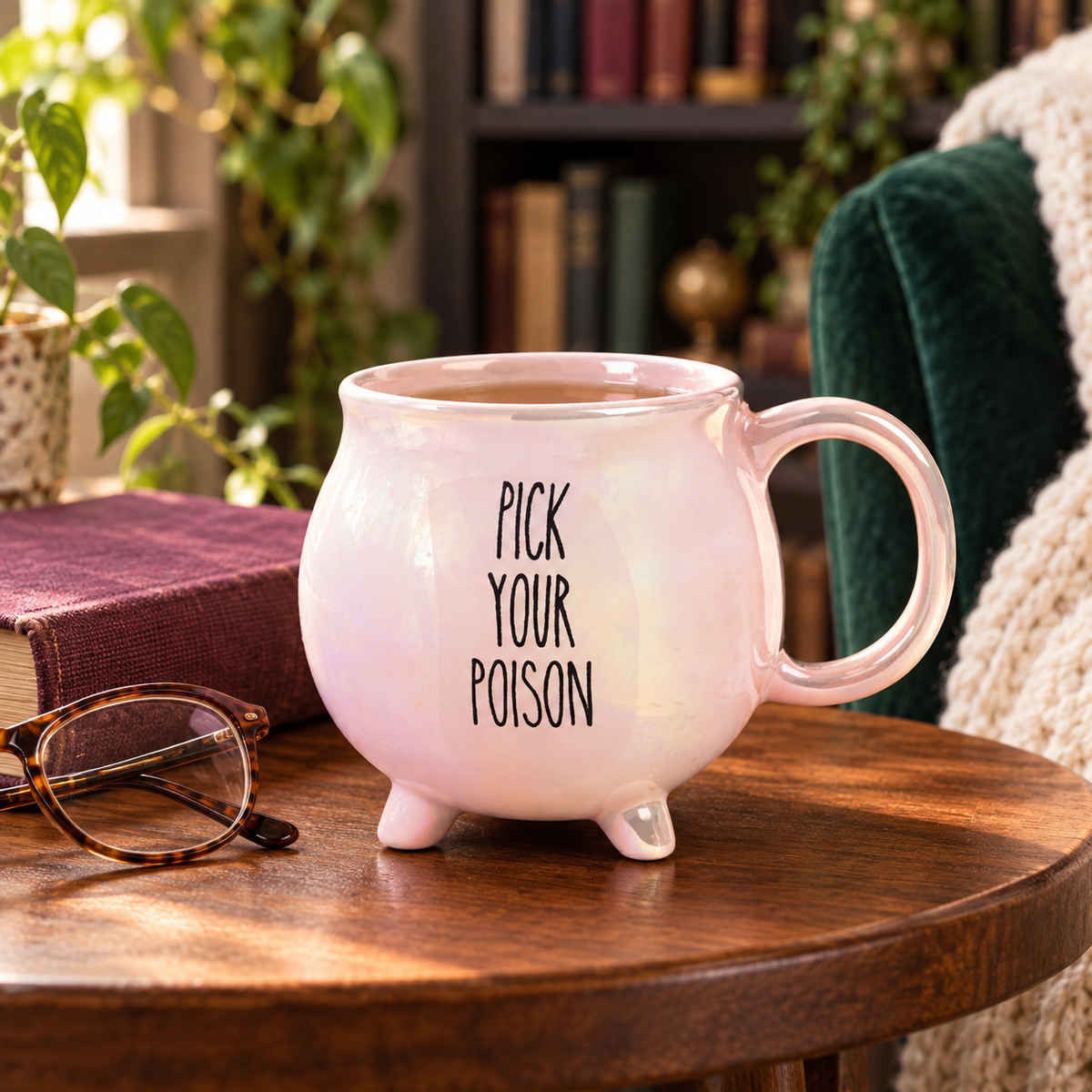 Pink iridescent Pick Your Poison cauldron mug on a wooden side table beside a book and reading glasses.