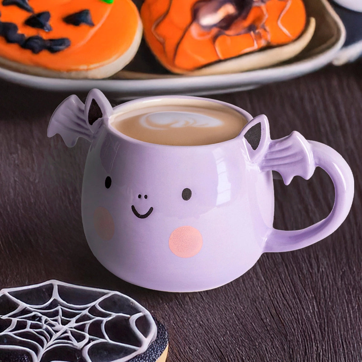 Angled view of a lilac bat mug on a dark wooden table beside decorated pumpkin and spiderweb treats.