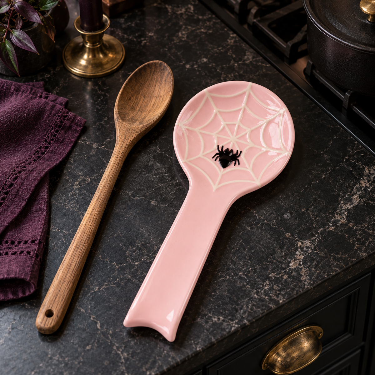 Pink spiderweb spoon rest with a black spider beside a wooden spoon and plum cloth on a dark kitchen counter.