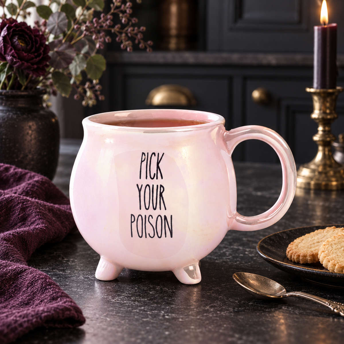 Pink iridescent Pick Your Poison cauldron mug on a dark counter beside cookies, plum linen and a lit candle.