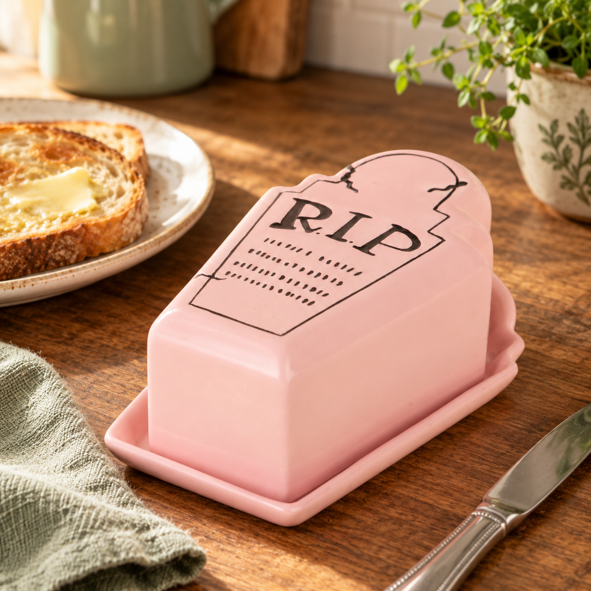 Pink R.I.P. tombstone butter dish beside buttered toast on a sunlit wooden kitchen counter.